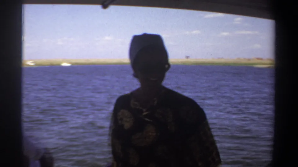A silhouetted woman in patterned clothing stands on the boat with the Nile and a distant Khartoum shoreline behind her.