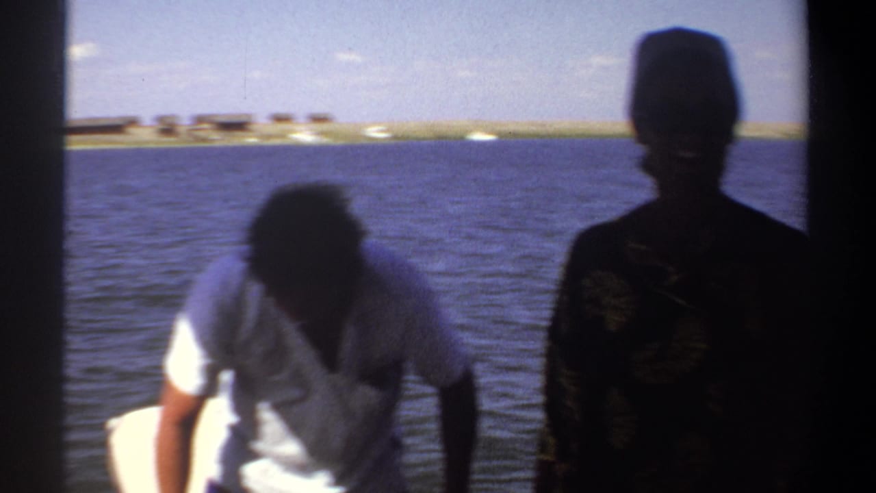1969 Khartoum home-movie frame showing two men on a small boat, one bent forward and one silhouetted, with open water behind them.