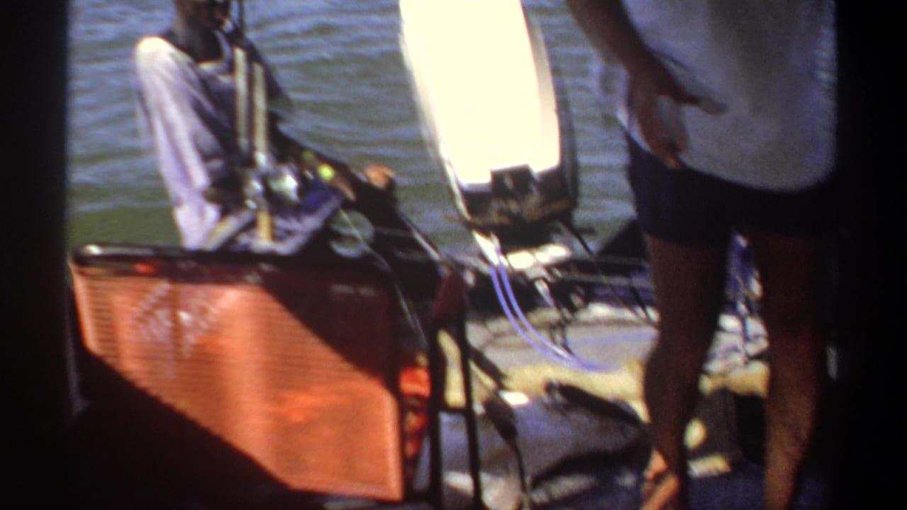 1969 Khartoum boating frame with a seated man near fishing rods and an outboard motor while another figure stands at the stern.