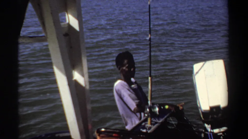 1969 Khartoum frame showing a man seated near fishing rods and an outboard motor, viewed through dark boat structure.