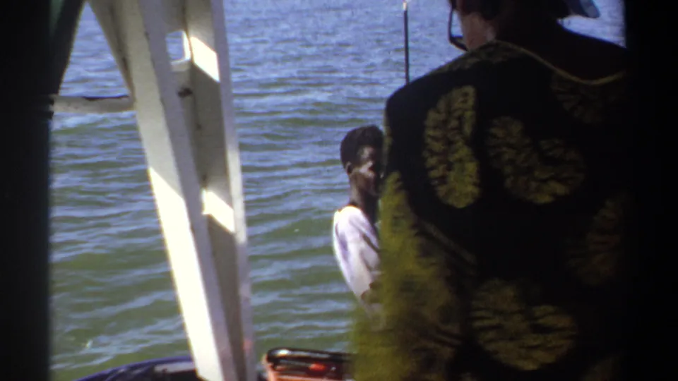 1969 Khartoum home-movie frame with a patterned-shirt figure in the foreground and a seated man near the motor on the water.