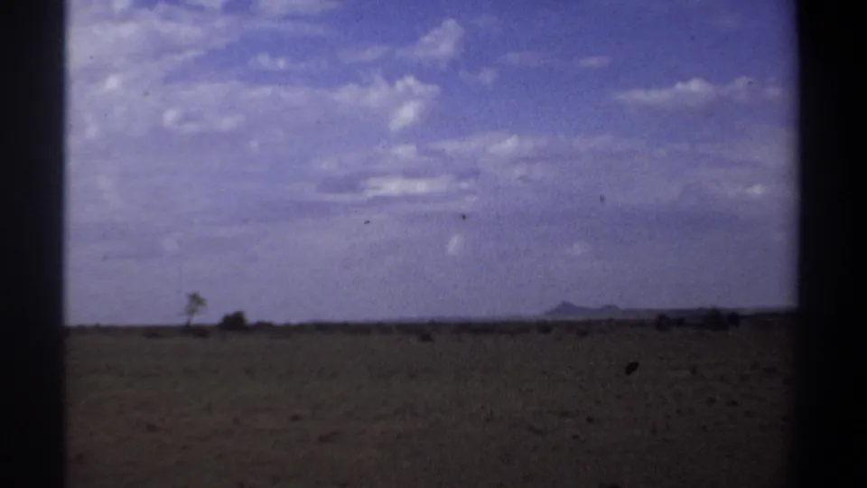 Wide 1969 Sudan landscape view with dry open ground, sparse vegetation, distant low hills, and a clouded blue sky.