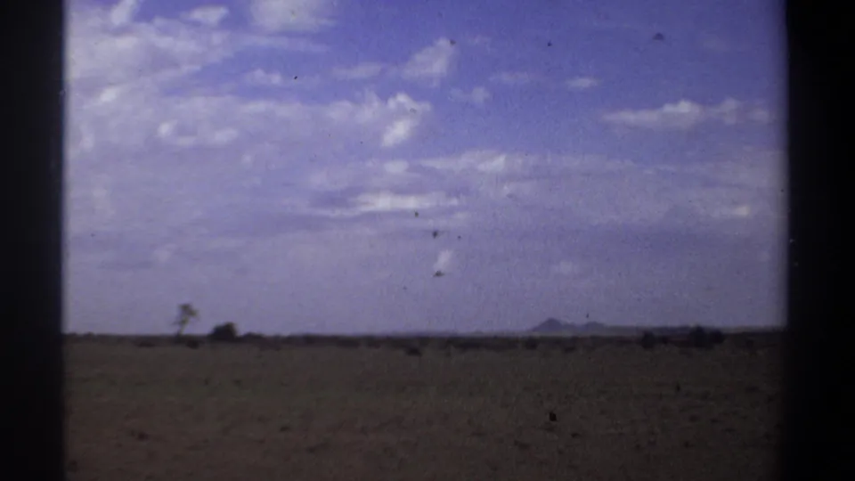 Wide Sudan travel-film frame showing open arid terrain, sparse trees, far hills, and a bright sky with scattered clouds.