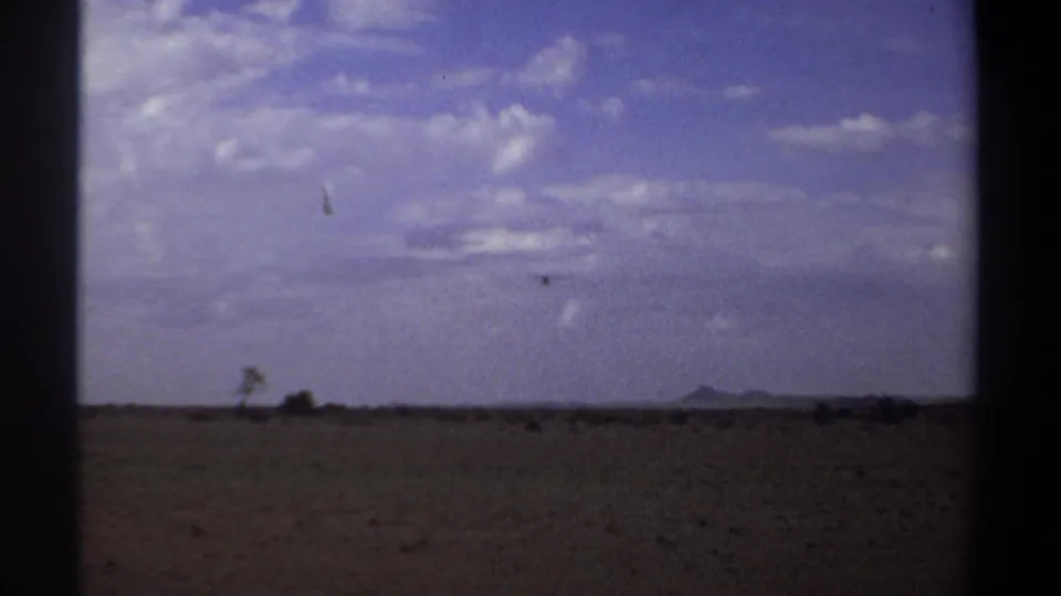 1969 Khartoum home-movie frame showing a small bird over a wide arid field with sparse vegetation and distant low hills.