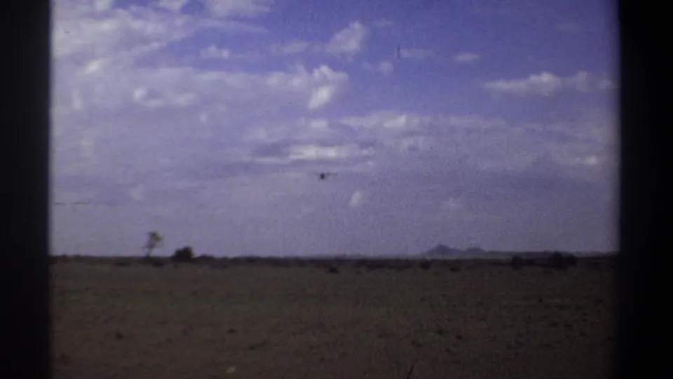 A 1969 Khartoum landscape frame with a distant bird above flat open ground, sparse plants, and a low hill on the horizon.