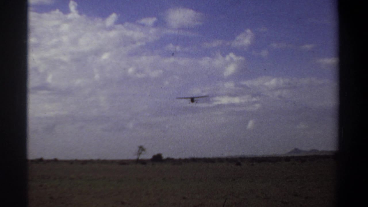 1969 Khartoum 8mm frame showing a small propeller aircraft distant over arid ground and a low horizon under scattered clouds.