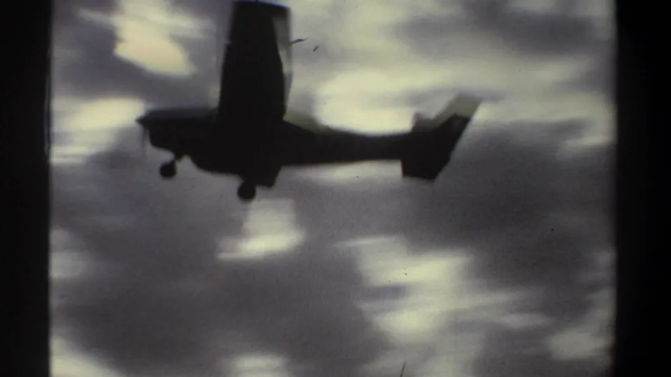 Silhouetted small aircraft passing overhead in 1969 Khartoum footage, with undercarriage visible against bright, soft-edged clouds.