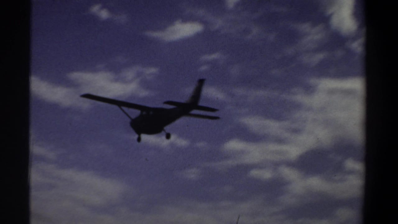 1969 Khartoum home-movie frame showing a small propeller airplane descending in silhouette under clouds.