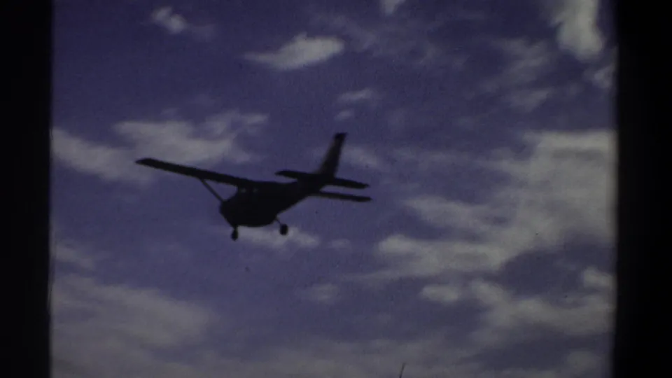 1969 Khartoum home-movie frame showing a small propeller airplane descending in silhouette under clouds.