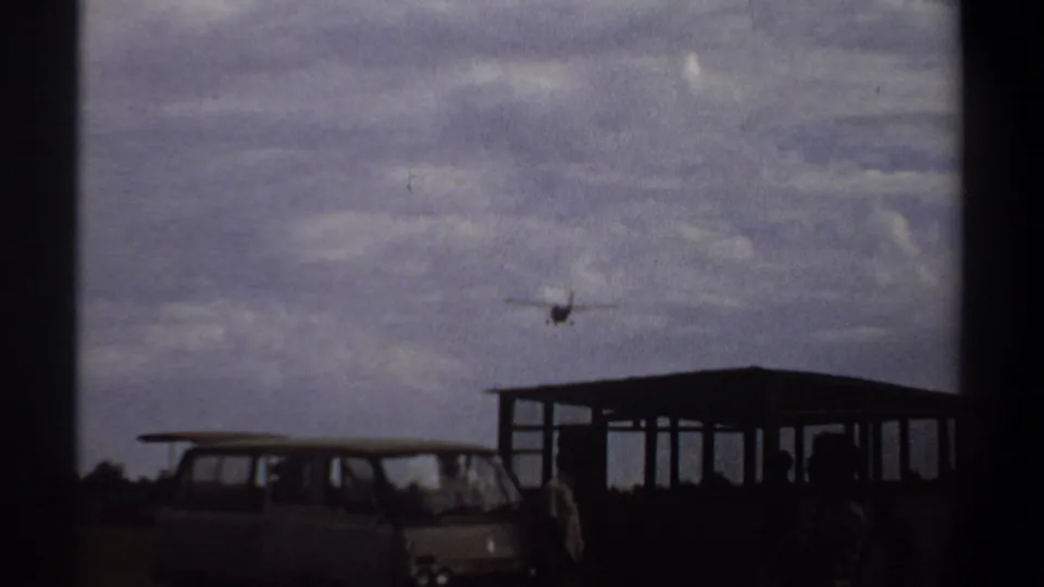 1969 Khartoum airport view with a van, open shelter, and distant descending aircraft in cloudy daylight.