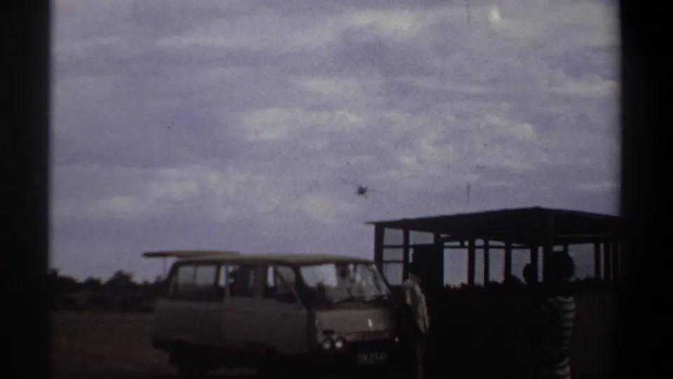 Khartoum 1969 airfield frame with a parked van, silhouetted people, shelter, and small aircraft in the distance.