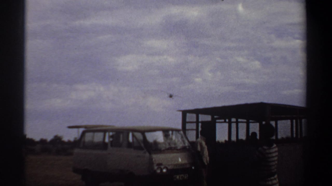 1969 Khartoum home-movie frame with a light van, open shelter, several onlookers, and a small propeller plane above the horizon.