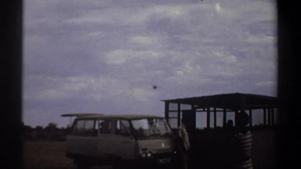 1969 Khartoum home-movie frame with a light van, open shelter, several onlookers, and a small propeller plane above the horizon.