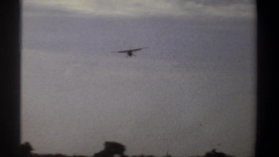 1969 Khartoum home-movie frame with the plane receding into the cloudy sky above a low, dark tree line.