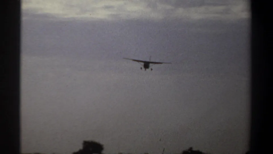 Distant propeller aircraft remains centered in the cloudy Khartoum sky, with landing gear visible in the 1969 8mm frame.