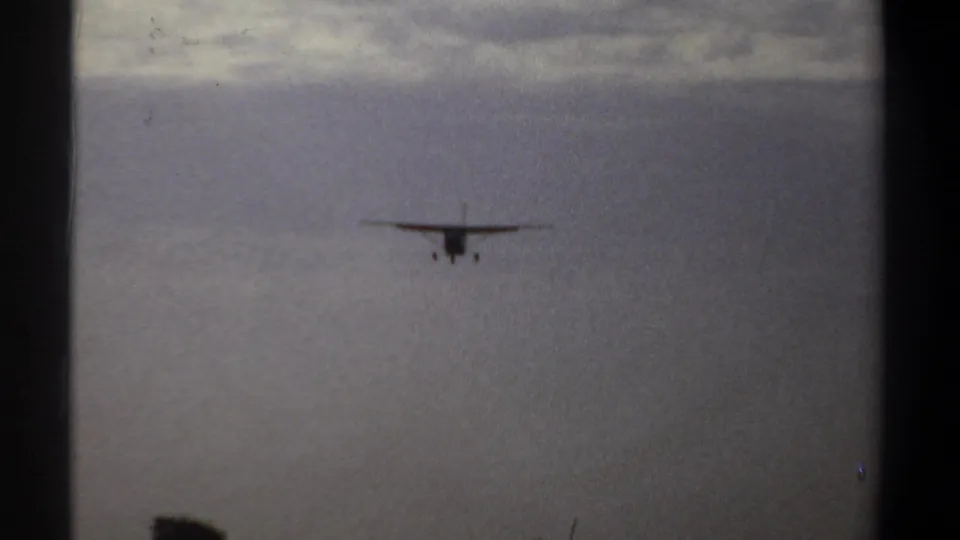 Propeller airplane descends through a hazy Khartoum sky, with dark tree silhouettes appearing along the lower edge of the 8mm frame.