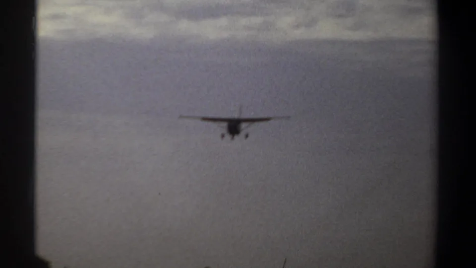 Small propeller airplane continues its 1969 approach over Khartoum, seen against soft cloud cover with faint trees below.