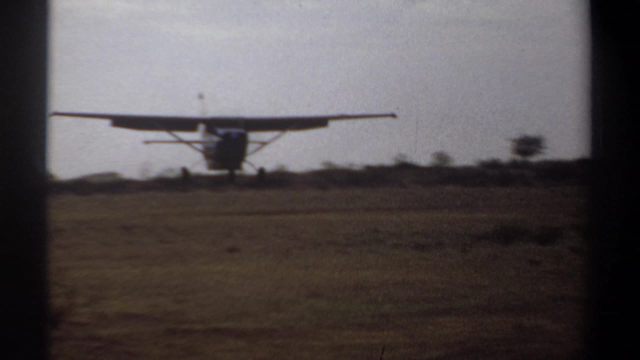 1969 Khartoum home-movie frame showing a small high-wing propeller aircraft approaching or landing on a grassy field.