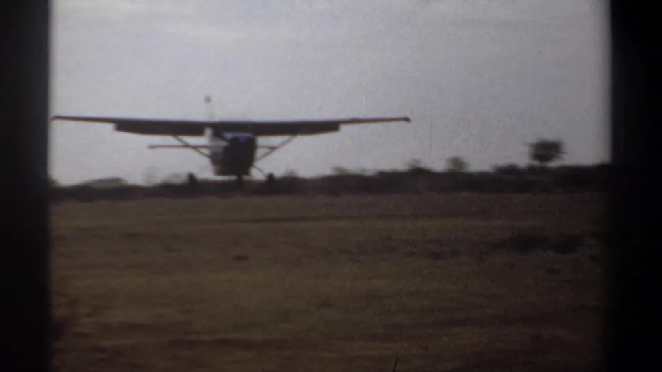 1969 Khartoum home-movie frame showing a small high-wing propeller aircraft approaching or landing on a grassy field.