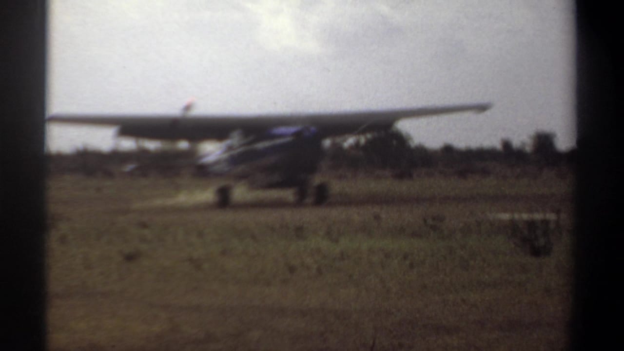 High-wing propeller aircraft on a grassy airstrip, viewed from the side at a distance.