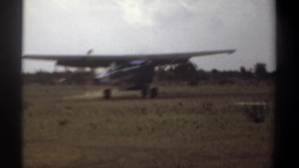 1969 Khartoum frame of a small blue and white propeller aircraft touching down or taxiing across a grass airstrip.