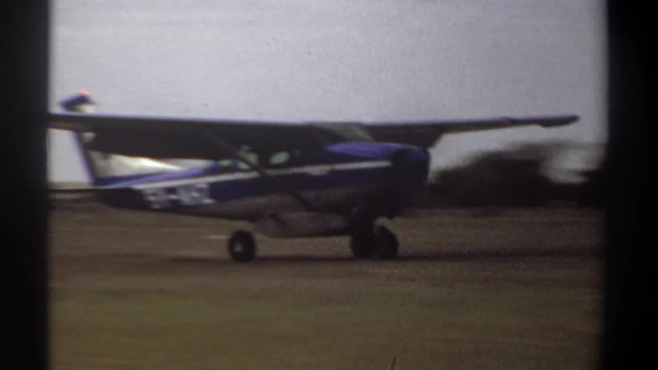 A blue and white high-wing aircraft taxis past the camera in 1969 Khartoum 8mm footage, with propeller blur visible.