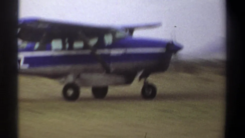 Close 1969 Khartoum home-movie view of the blue and white high-wing aircraft taxiing on grass with its propeller spinning.