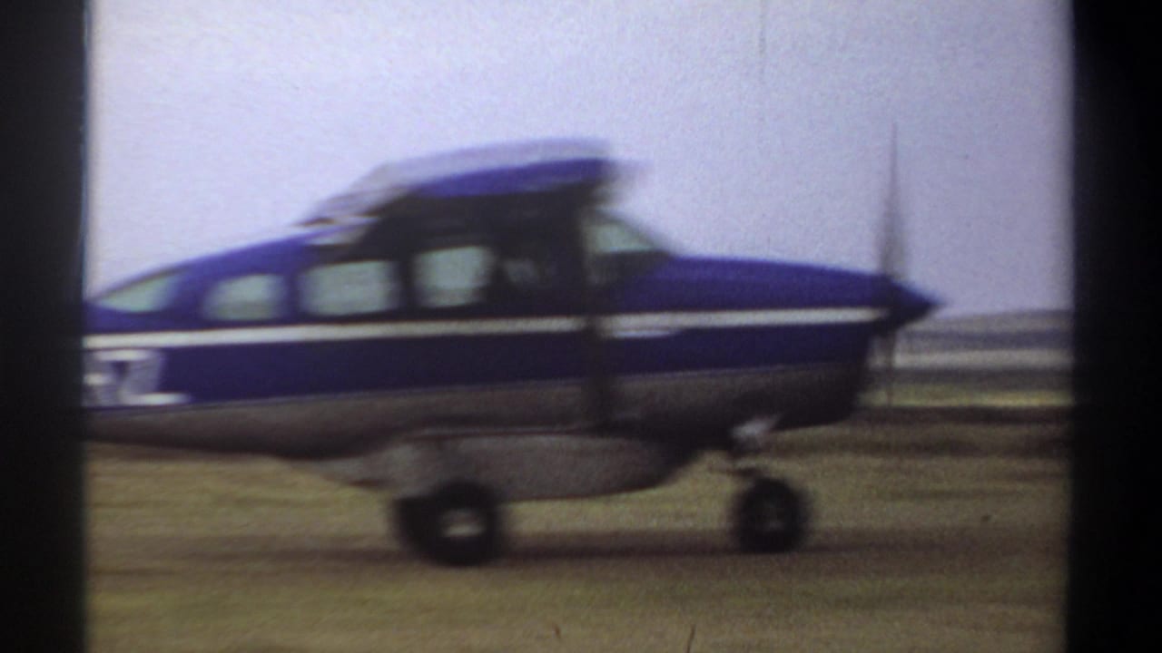 A blue and white single-engine airplane passes close to the camera on a grass airstrip in 1969 Khartoum home-movie footage.
