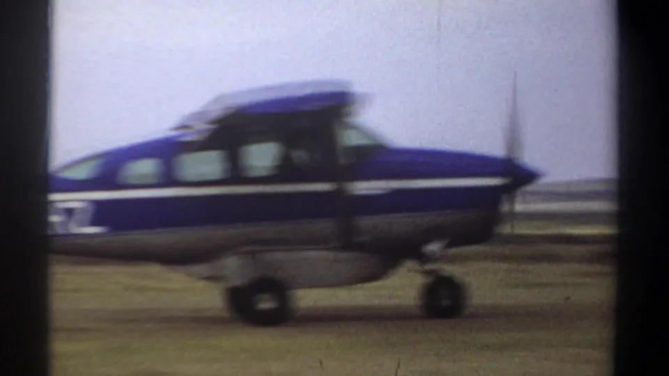A blue and white single-engine airplane passes close to the camera on a grass airstrip in 1969 Khartoum home-movie footage.