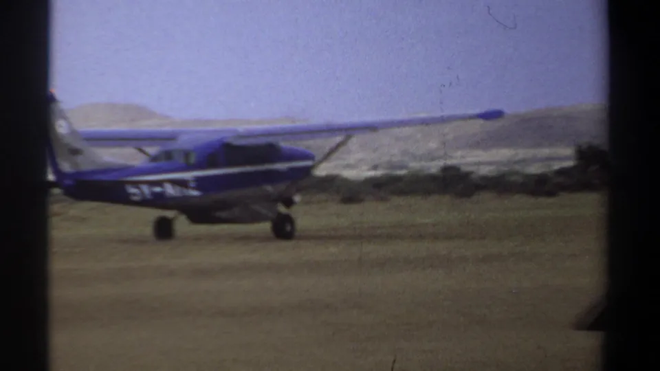 The aircraft continues across the grass airstrip in 1969 Sudan footage, seen against pale sky and distant low hills.