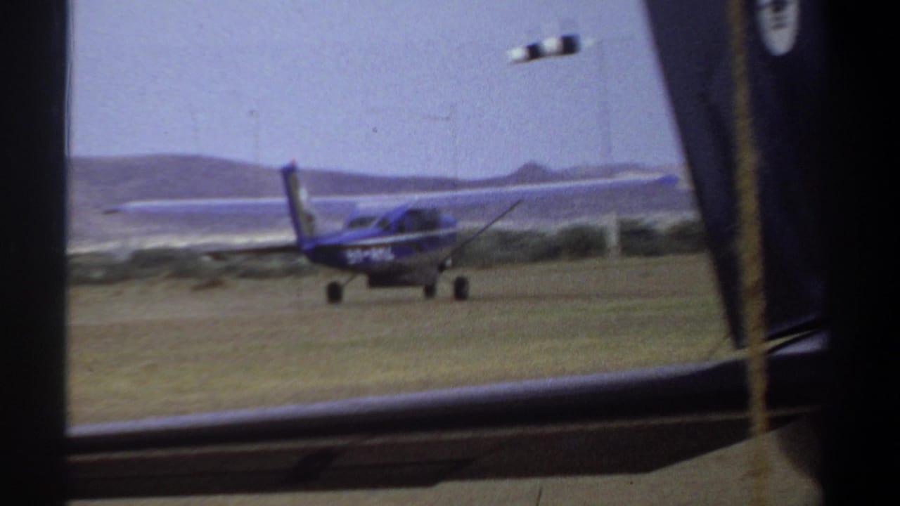 1969 Khartoum home-movie frame showing a small blue and white single-engine airplane on grass with distant hills and film borders.