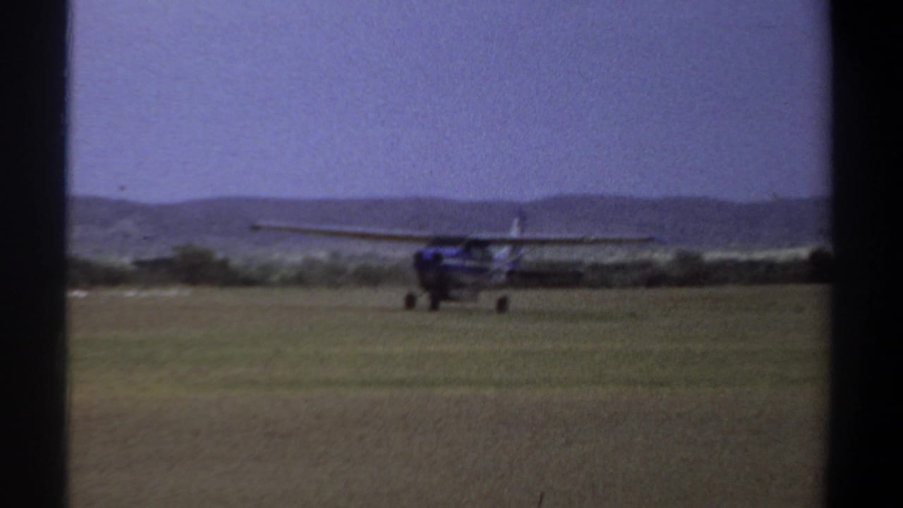 Small airplane taxiing across a grassy field with low hills in the background.