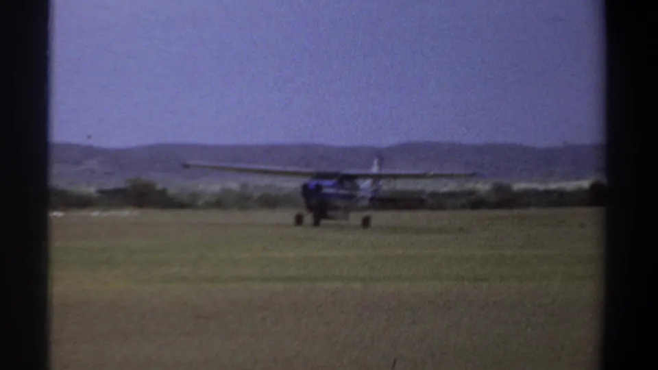 1969 Khartoum frame of a small blue and white aircraft moving across a grassy airfield under a hazy sky.