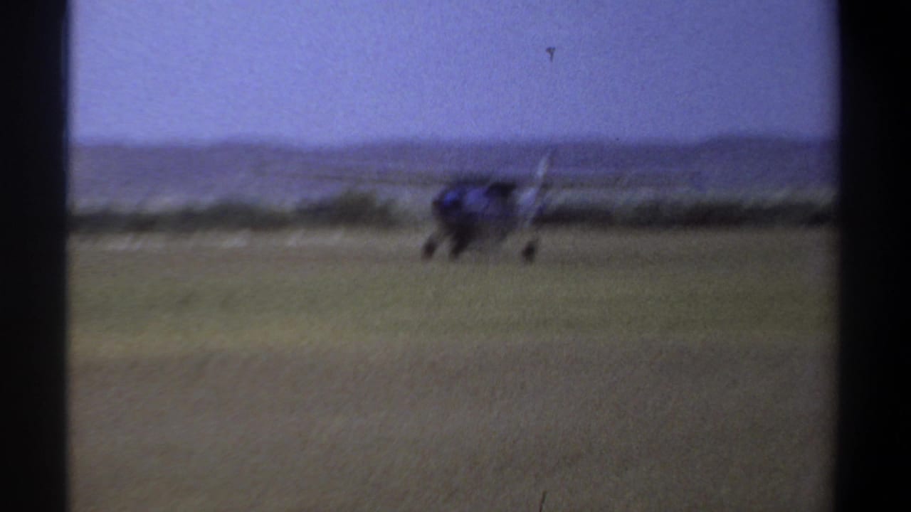 1969 Khartoum home-movie frame showing a small blue aircraft on a grassy airfield, seen through dark film-frame borders.