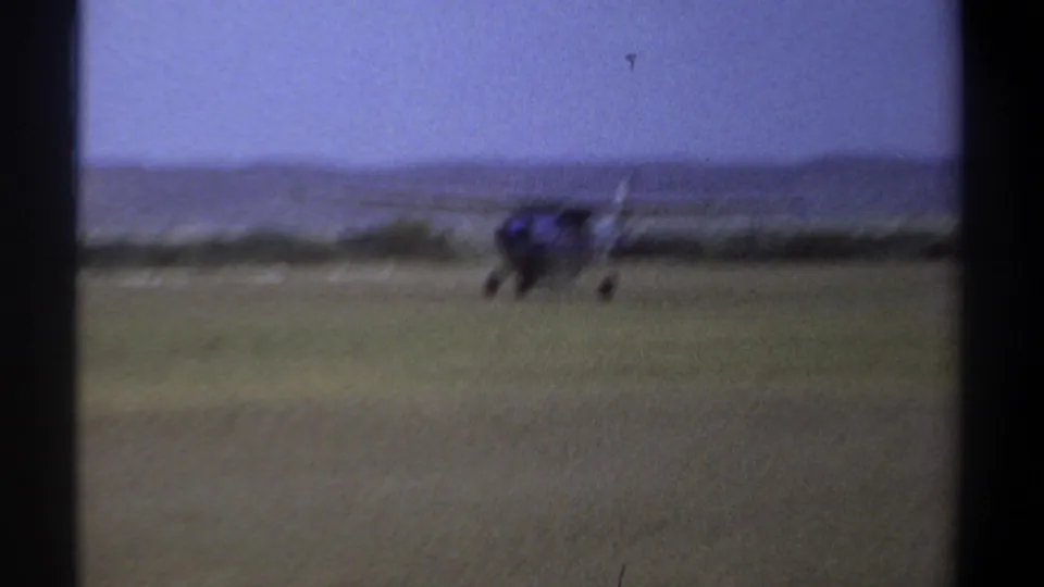 1969 Khartoum home-movie frame showing a small blue aircraft on a grassy airfield, seen through dark film-frame borders.