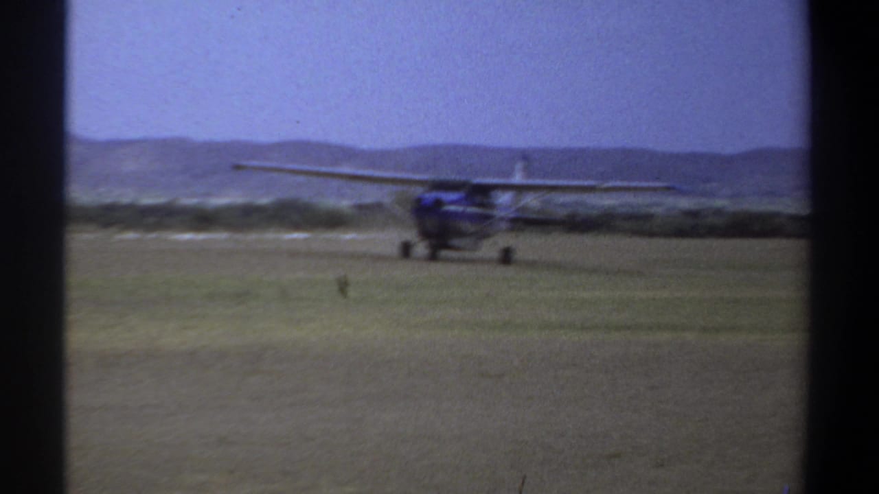 Blue high-wing plane taxiing across open grass with low hills behind it.