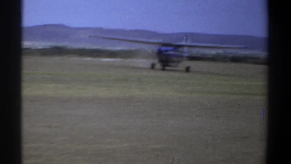 Blue single-engine aircraft crossing a grassy airfield in the 1969 Khartoum footage, partly softened by 8mm motion blur.