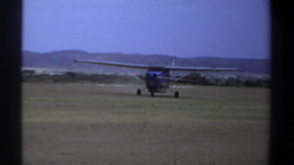 Final frame of the 1969 Khartoum airfield scene, with the small blue aircraft centered against grass, scrub, hills, and sky.