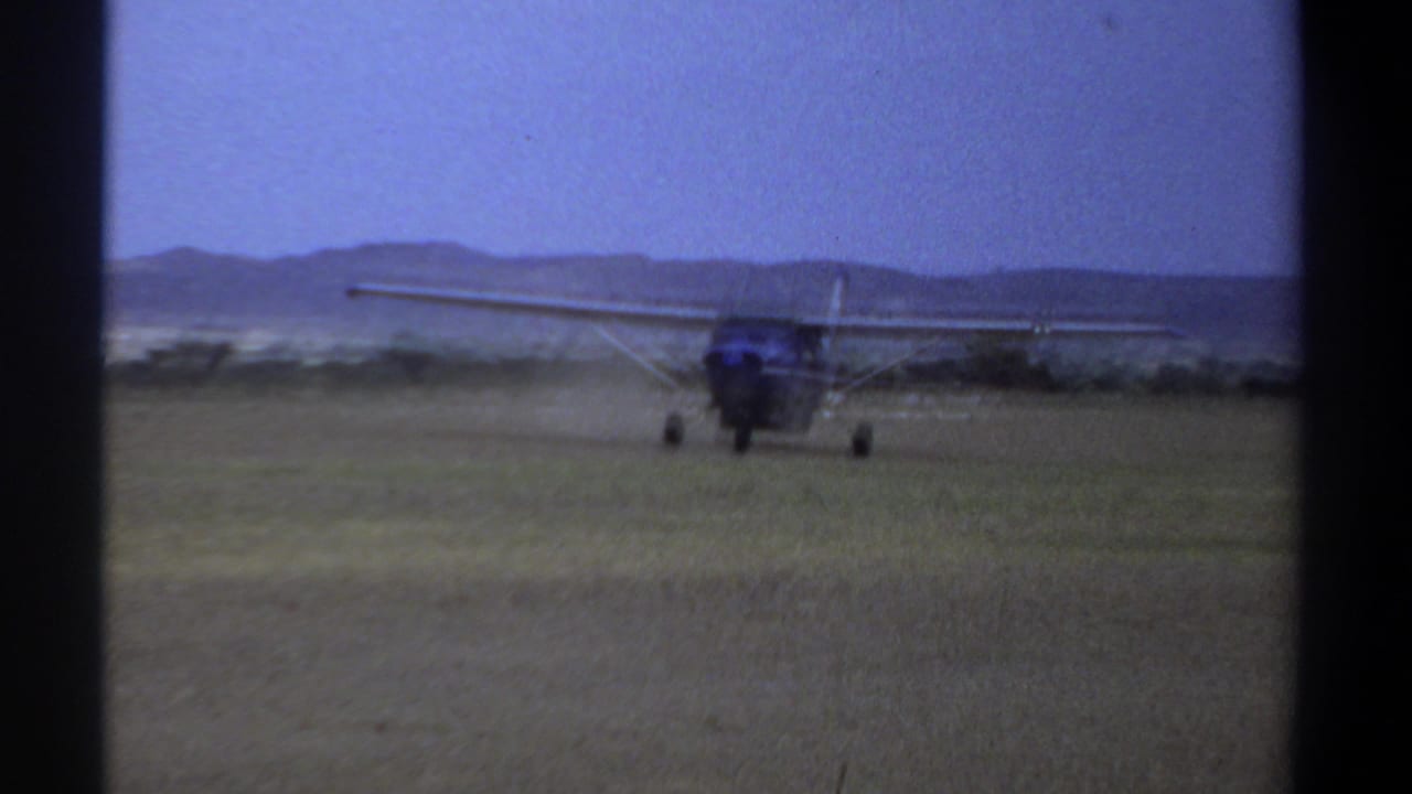 1969 Khartoum home-movie frame showing a small high-wing propeller airplane taxiing across a grass airfield with low hills in the background.