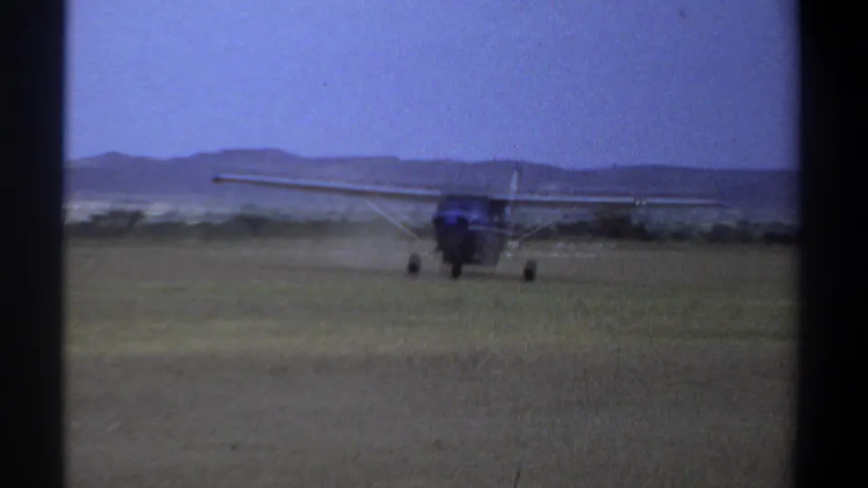 1969 Khartoum home-movie frame showing a small high-wing propeller airplane taxiing across a grass airfield with low hills in the background.