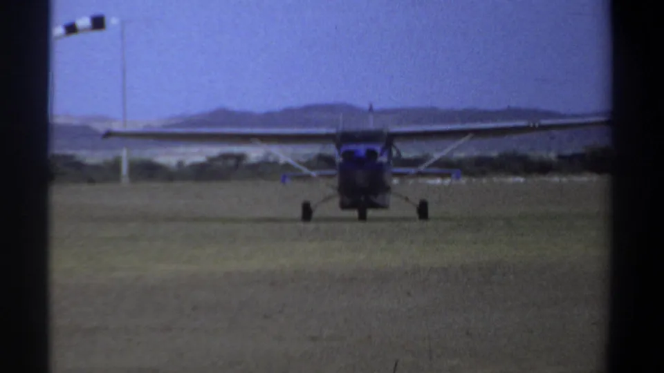 1969 Khartoum frame of a small high-wing propeller airplane centered on a grass airstrip, with distant hills under a pale blue sky.