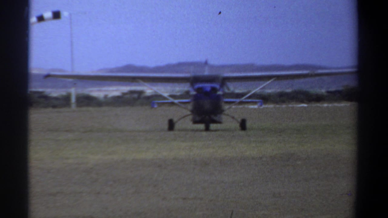 1969 Khartoum home-movie frame showing a small high-wing propeller plane taxiing toward the camera on a grass strip.