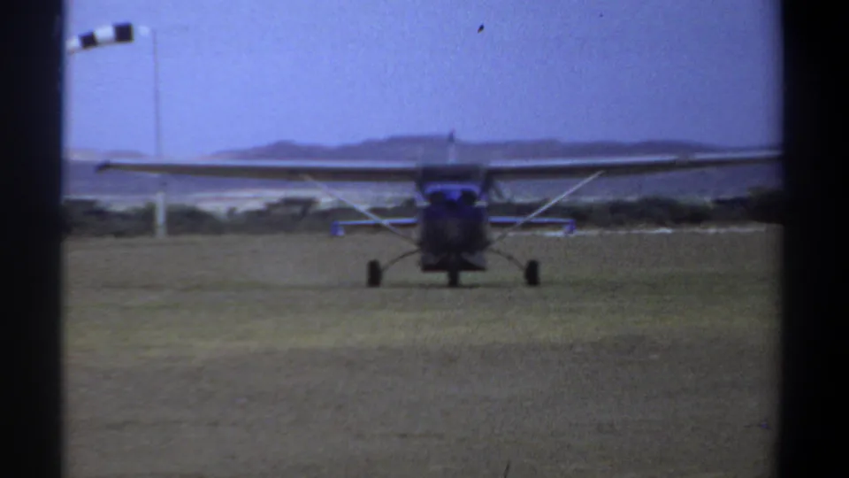 1969 Khartoum home-movie frame showing a small high-wing propeller plane taxiing toward the camera on a grass strip.