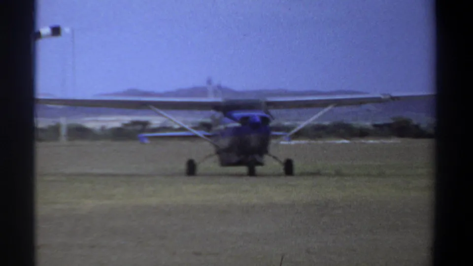 1969 Khartoum frame of a blue and white single-engine plane moving on a grass airfield under a hazy sky.