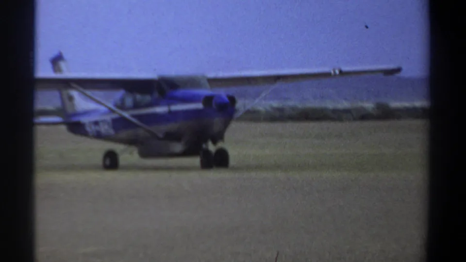 1969 Khartoum 8mm frame showing a side view of a small blue and white propeller plane on a grass airfield.