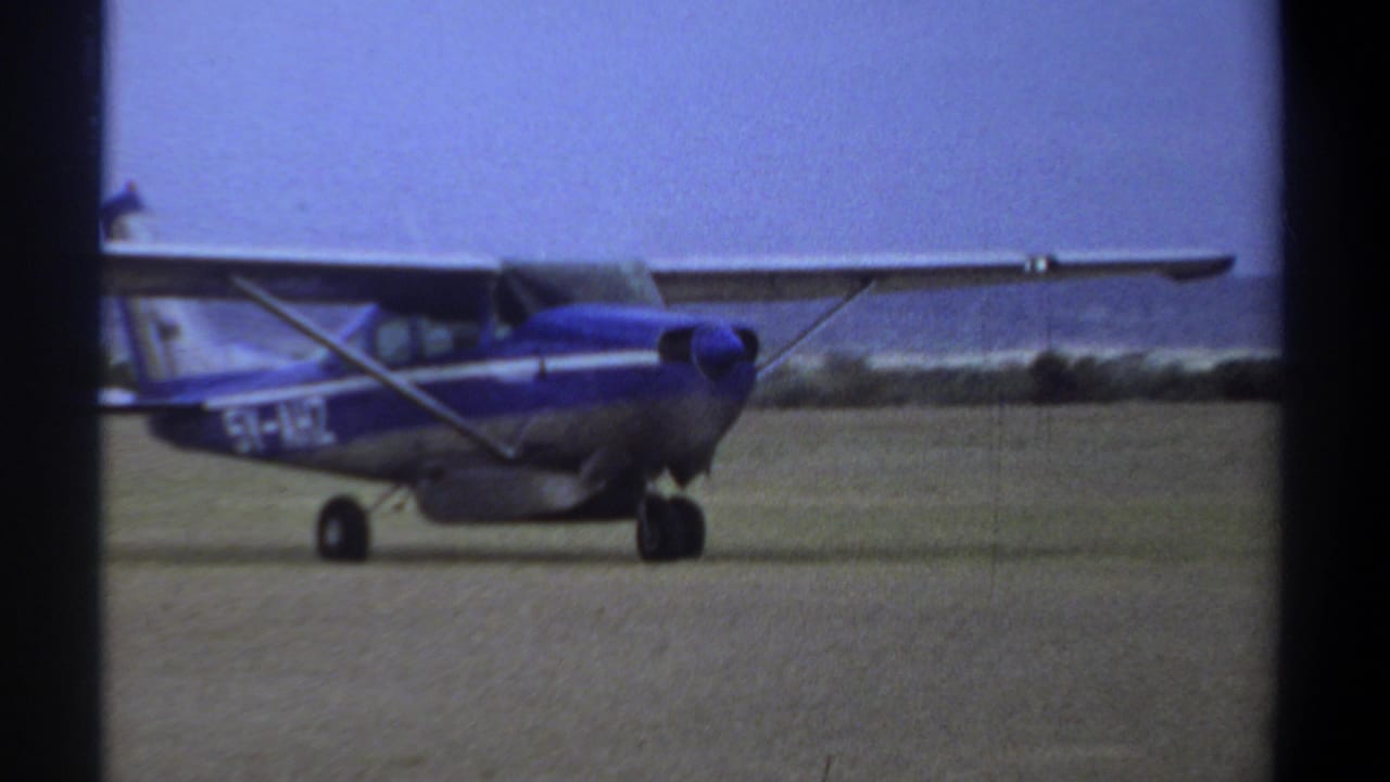 Blue and silver light aircraft taxiing on a dirt runway in 1969 Khartoum footage, seen from a handheld 8mm viewpoint.