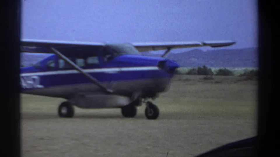 Closer 8mm frame of the blue and silver aircraft in Khartoum, showing the wing strut, front landing gear, and partial registration lettering.