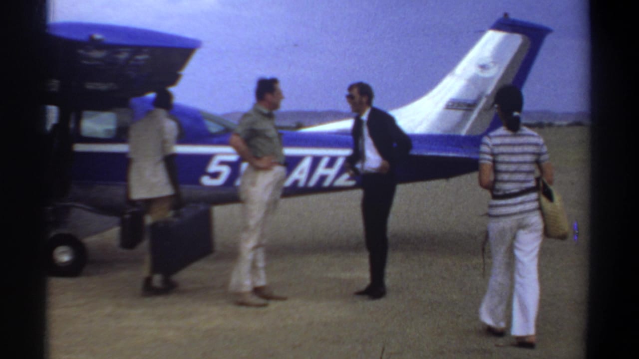 1969 Khartoum airfield frame with adults and luggage gathered beside a blue-and-white small aircraft.