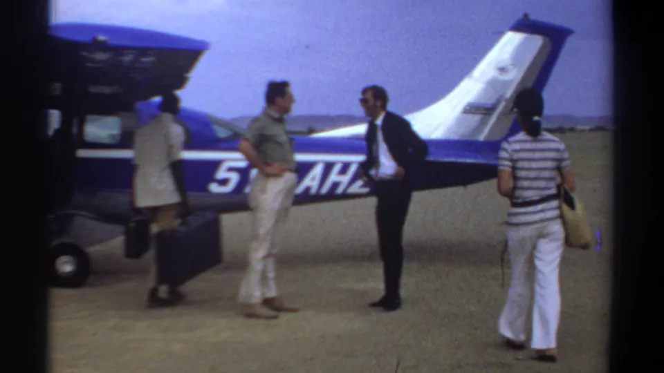 1969 Khartoum airfield frame with adults and luggage gathered beside a blue-and-white small aircraft.