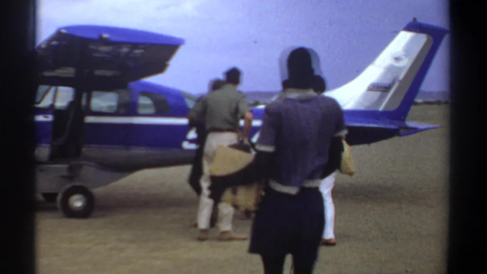 1969 Khartoum home-movie frame with passengers carrying bags toward a small aircraft on an airfield.